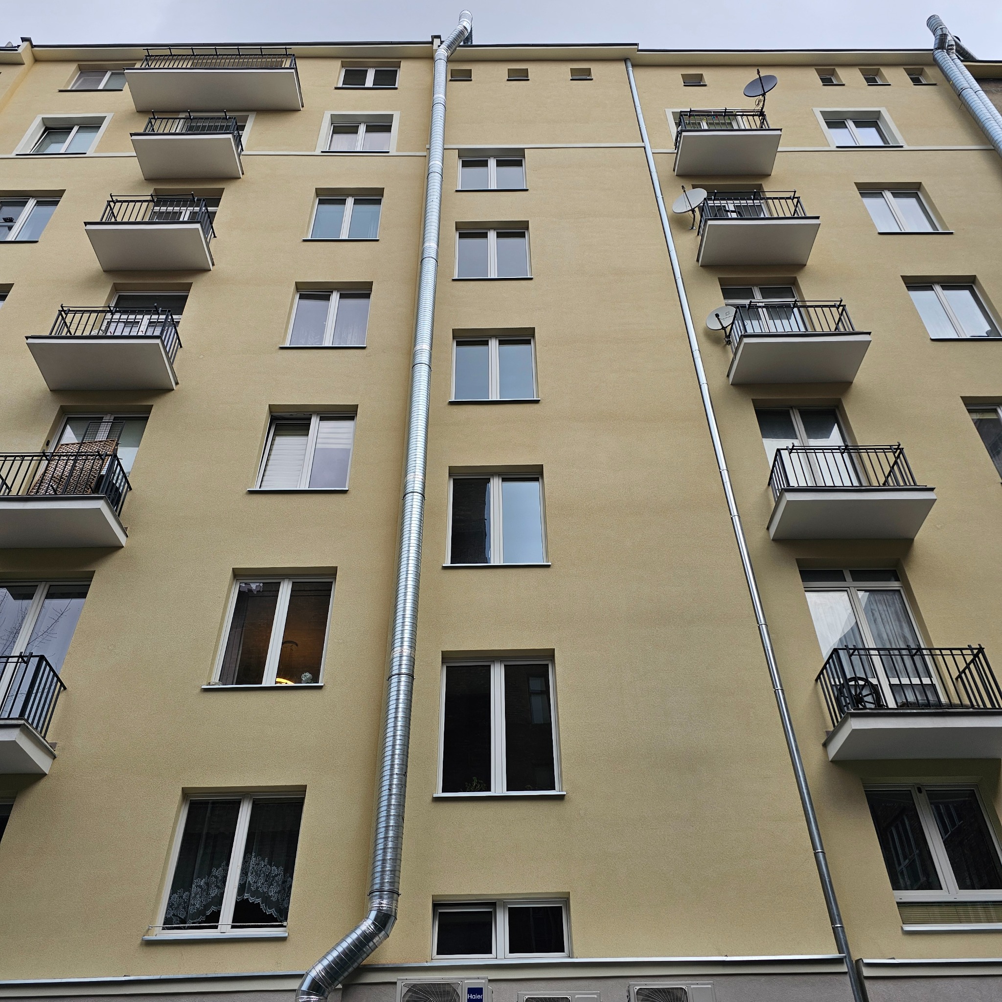 Elewacja budynku mieszkalnego z zainstalowanymi zewnętrznymi kanałami wentylacyjnymi, widok z dołu na balkony z czarnymi balustradami i okna z białymi ramami.