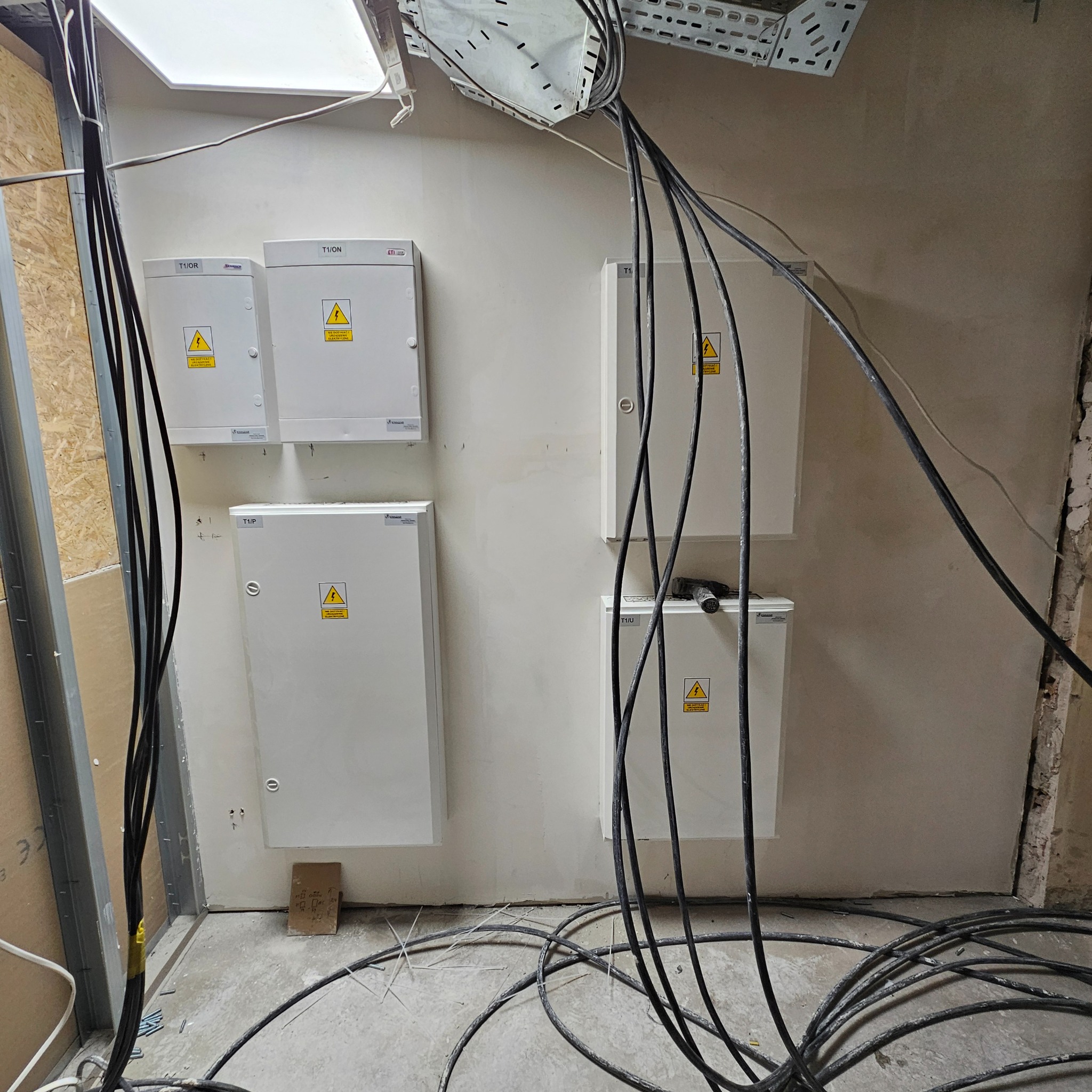 Rozdzielnice elektryczne T1/ON i T1/OR z żółtymi naklejkami ostrzegawczymi, liczne kable zwisające ze ściany w surowym pomieszczeniu podczas instalacji.