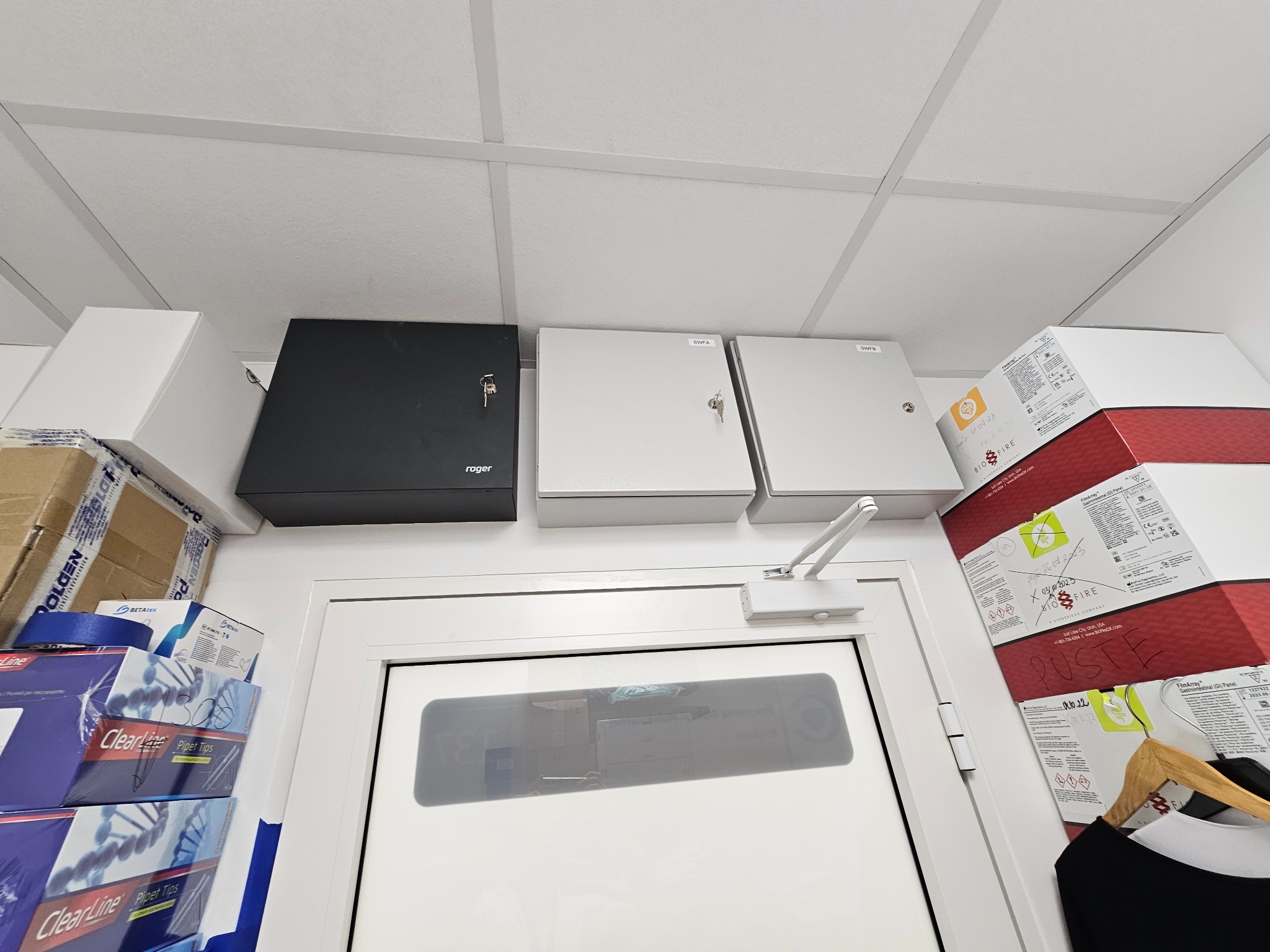 Szafki sterownicze Roger i SWFA zamontowane pod sufitem, widoczne kluczyki w zamkach, obok drzwi z samozamykaczem oraz kartony z materiałami laboratoryjnymi.