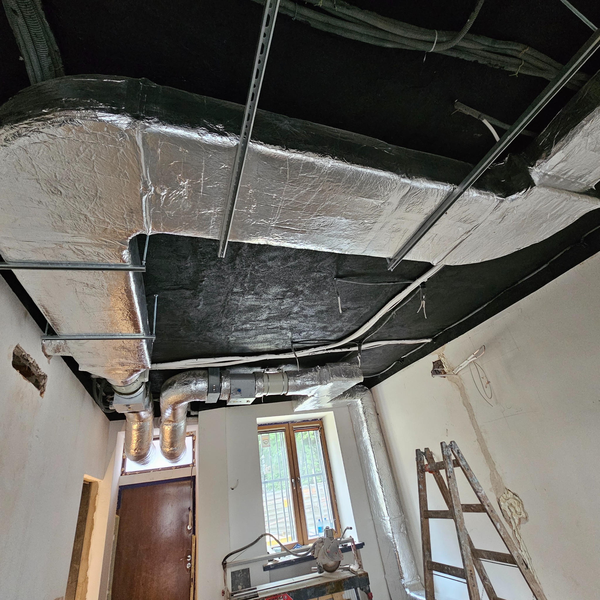 Widok z dołu na montowane przewody wentylacyjne z izolacją aluminiową, podwieszone pod czarnym sufitem, z widocznymi przewodami elektrycznymi, fragmentem okna i drabiną w surowym wnętrzu.