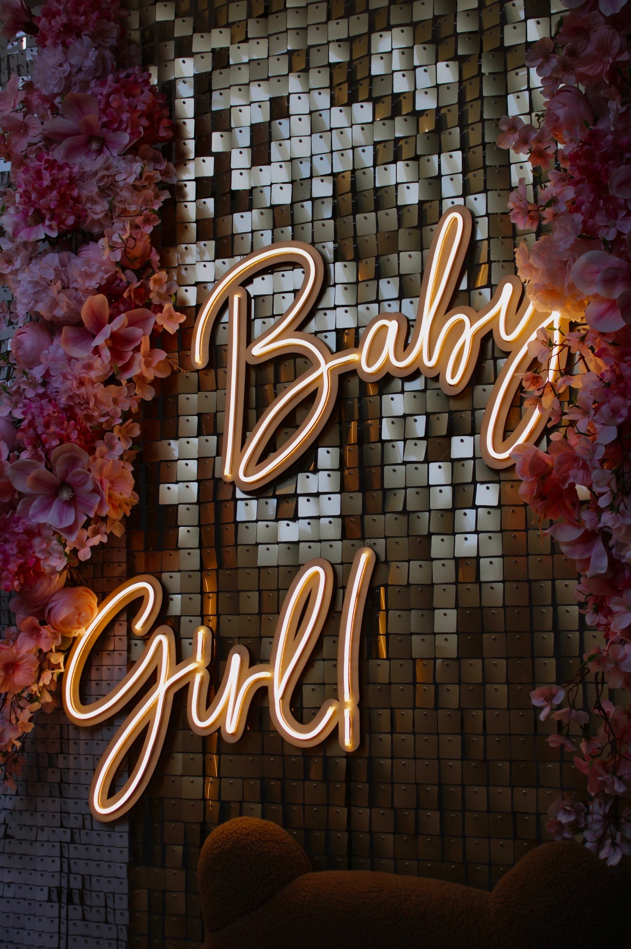 Neonowy napis 'Baby Girl' na błyszczącym złotym tle z kwadratowych cekinów, otoczony różowymi kwiatami, aranżacja dekoracyjna.