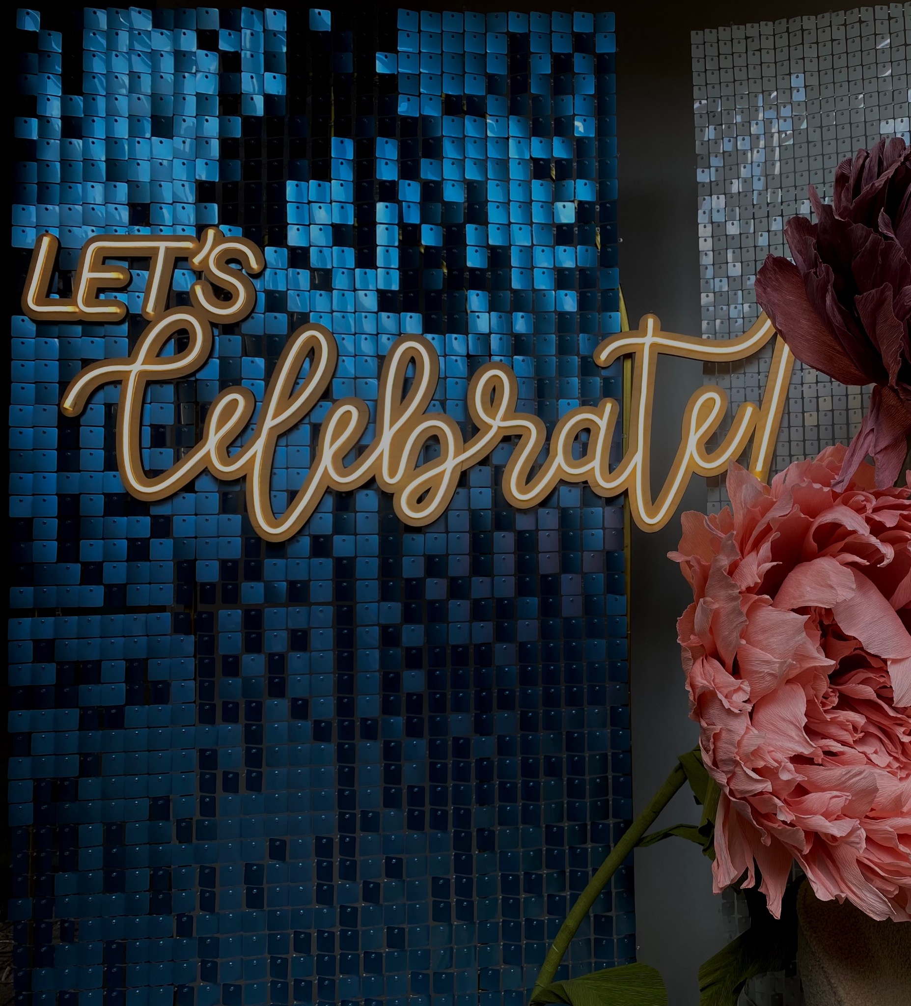 Błyszcząca ścianka z niebieskich kwadratów z napisem neonowym 'Let's Celebrate' i dekoracyjnymi papierowymi kwiatami w odcieniach różu i bordo.