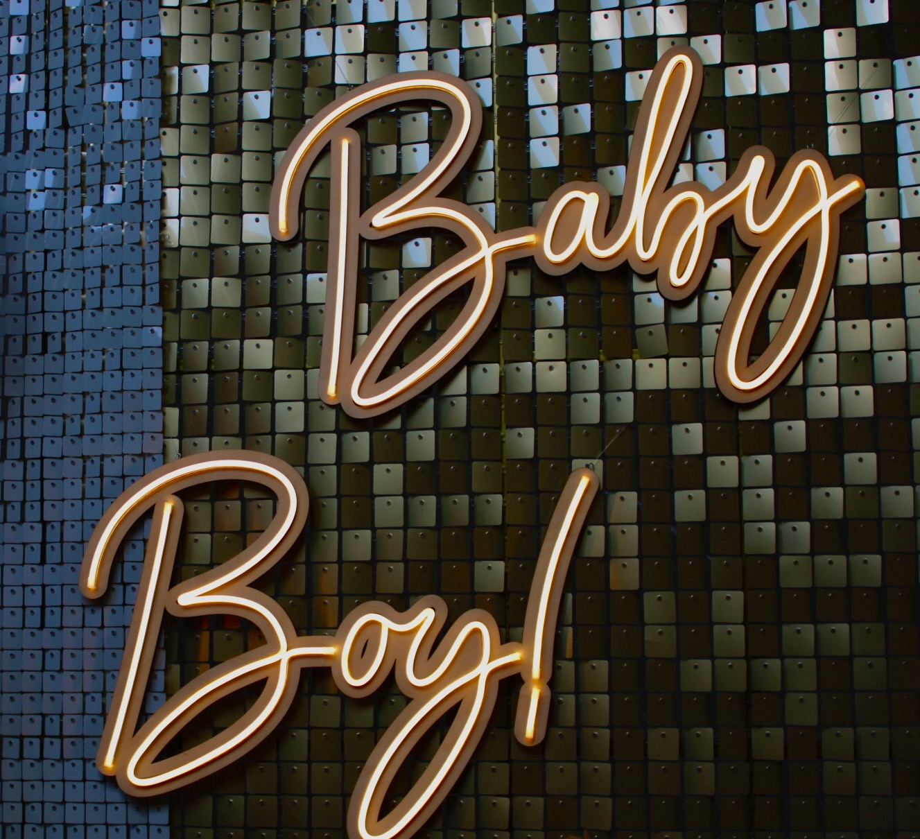 Świecący neonowy napis 'Baby Boy!' na błyszczącym, kwadratowym tle w odcieniach szarości i czerni.