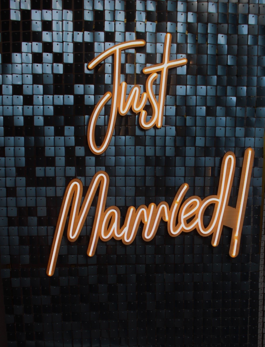 Dekoracja ścienna z błyszczących kwadratów i neonowym napisem 'Just Married' w kolorze pomarańczowym z białą obwódką.