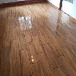 Rys bud - Świeżo ułożona, błyszcząca podłoga z płytek imitujących drewno, ułożonych w prostokątnym wzorze, z widocznymi rurami grzewczymi pod sufitem i fragmentem ościeżnicy drzwi.