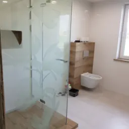 Nowoczesna łazienka z kabiną prysznicową ze szkła z motywem kwiatowym, podwieszaną toaletą i drewnopodobną zabudową stelaża, białe płytki na ścianach i podłodze, widoczne narzędzia i chemia budowlana.