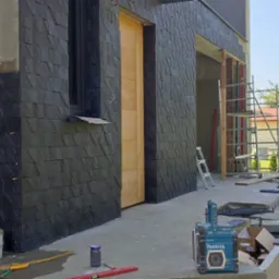 Częściowo wykończona elewacja budynku z czarnej, łupkowej płytki, widoczne narzędzia glazurnicze, drabina i rusztowanie.