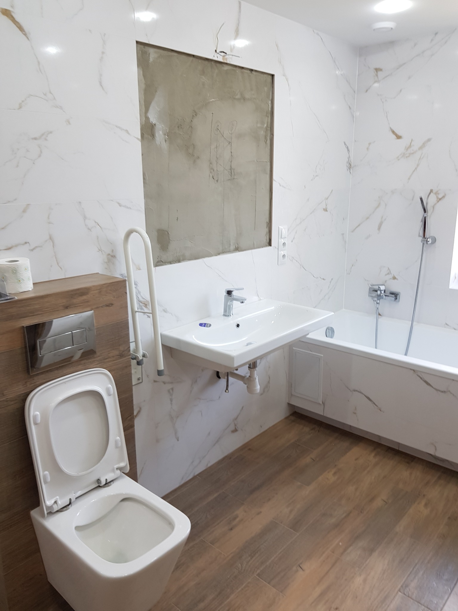 Częściowo wykończona łazienka z biało-złotymi marmurowymi płytkami ściennymi, drewnopodobną podłogą, wiszącą toaletą, umywalką i wanną; widoczne niedokończone miejsce na lustro.