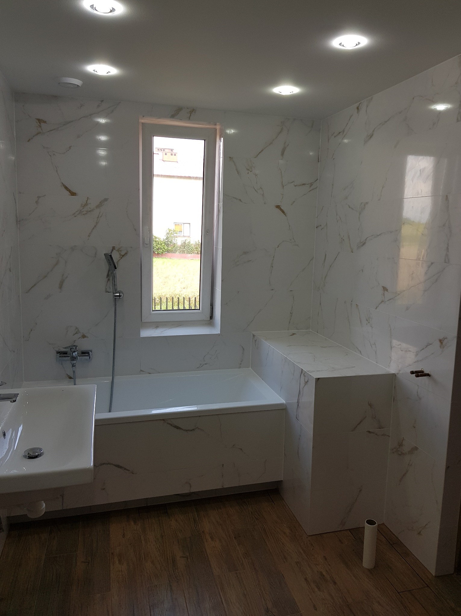 Nowoczesna łazienka z wanną, umywalką i zabudową, wykończona białymi płytkami imitującymi marmur z delikatnymi złotymi żyłkami, drewnopodobna podłoga, okno z widokiem na zieleń.