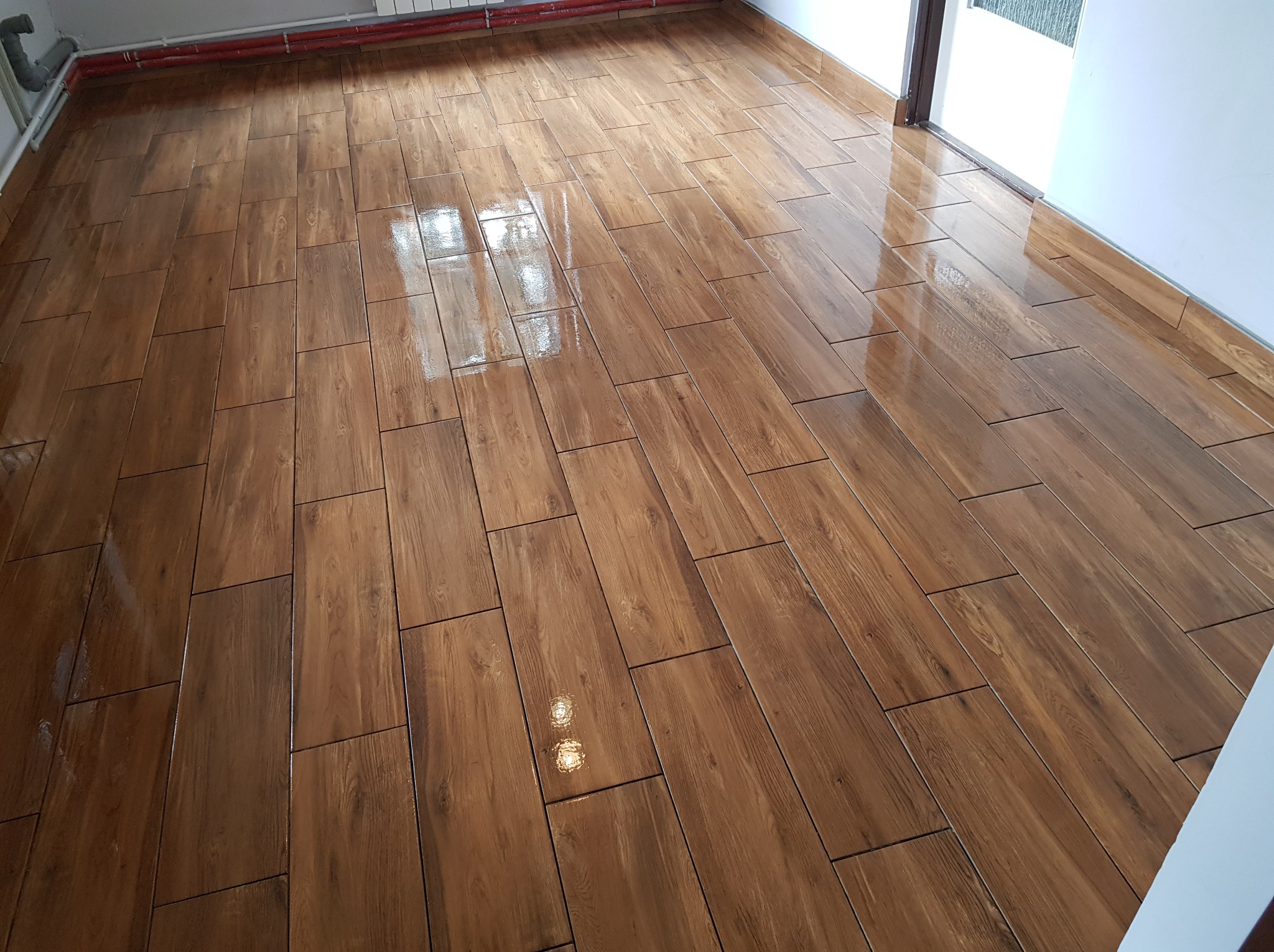 Świeżo ułożona, błyszcząca podłoga z płytek imitujących drewno, ułożonych w prostokątnym wzorze, z widocznymi rurami grzewczymi pod sufitem i fragmentem ościeżnicy drzwi.