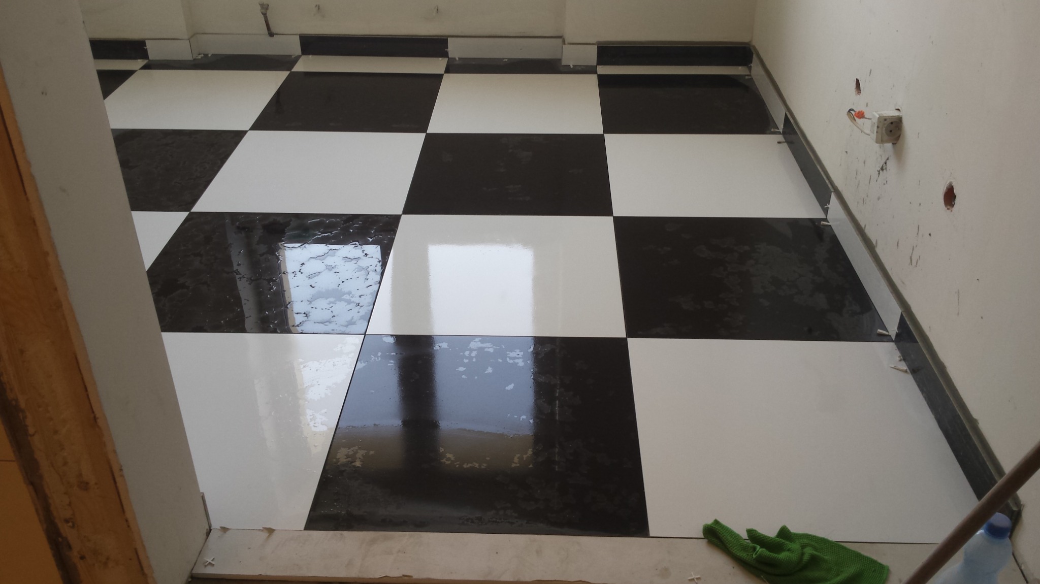 Ułożona podłoga w szachownicę z czarno-białych płytek, widoczne krzyżyki dystansowe, ślady fugowania na czarnych płytkach i zielona ścierka w rogu pomieszczenia.