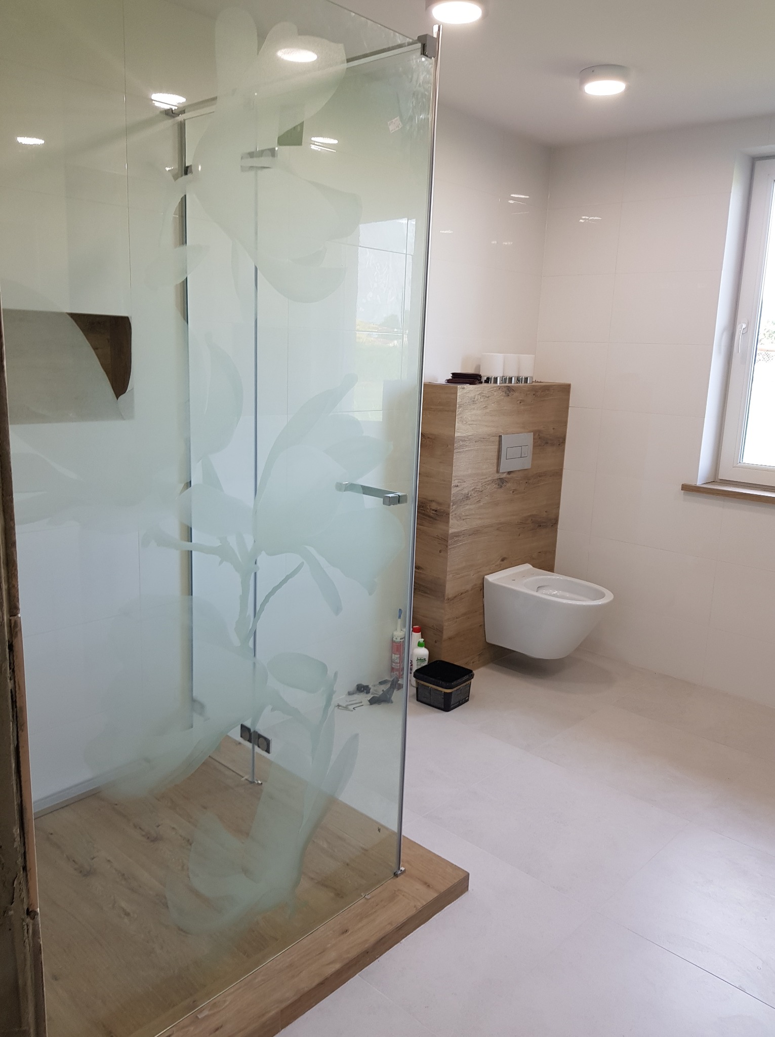 Nowoczesna łazienka z kabiną prysznicową ze szkła z motywem kwiatowym, podwieszaną toaletą i drewnopodobną zabudową stelaża, białe płytki na ścianach i podłodze, widoczne narzędzia i chemia budowlana.