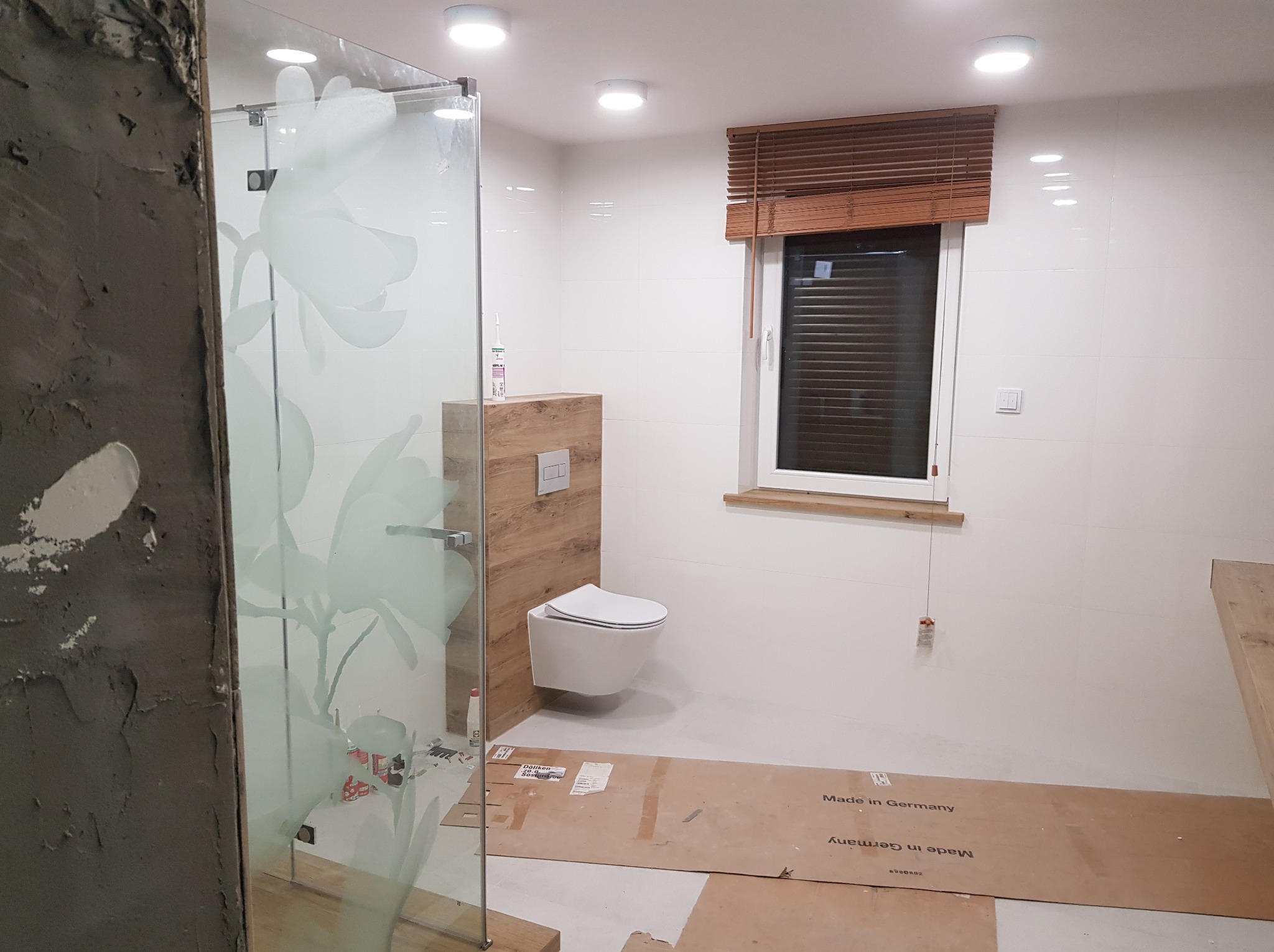 Wnętrze łazienki w trakcie prac wykończeniowych: kabina prysznicowa z wzorem kwiatowym, podwieszana toaleta na tle drewnopodobnej zabudowy, białe płytki ścienne i brązowe żaluzje okienne, podłoga...