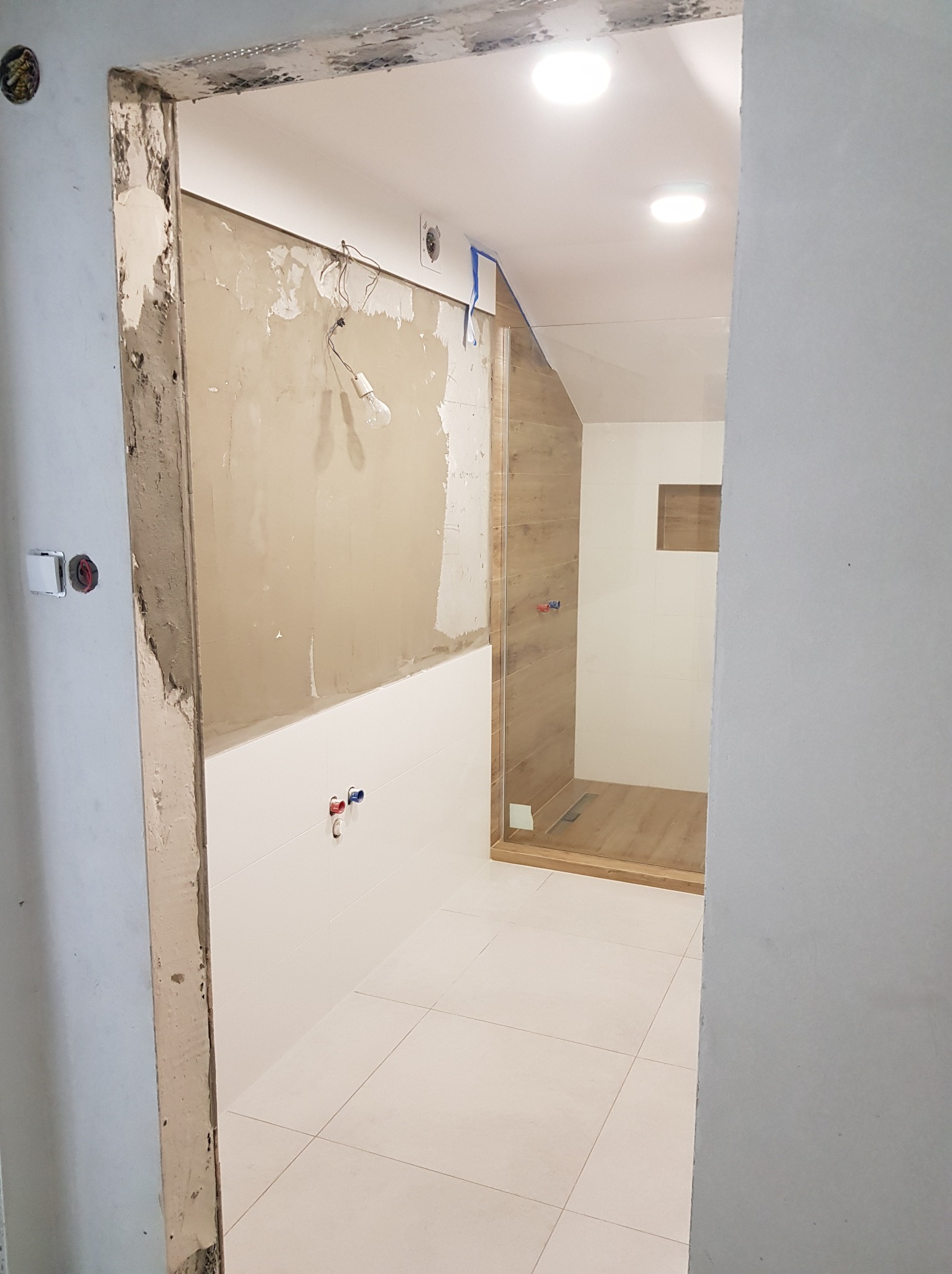 Wnętrze łazienki w trakcie prac wykończeniowych, widoczne surowe ściany, białe płytki podłogowe, częściowo wykończona kabina prysznicowa z drewnopodobnymi panelami i szklaną ścianą, oraz wisząca...