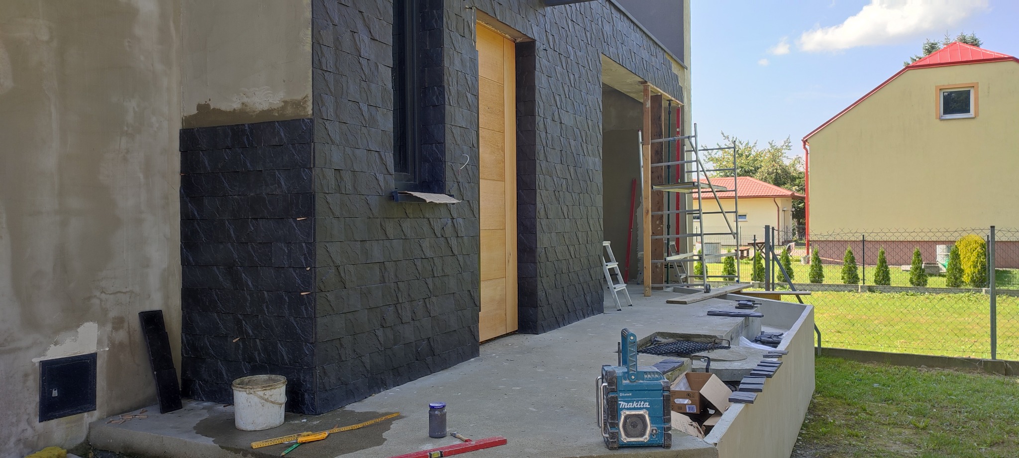 Częściowo wykończona elewacja budynku z czarnej, łupkowej płytki, widoczne narzędzia glazurnicze, drabina i rusztowanie.