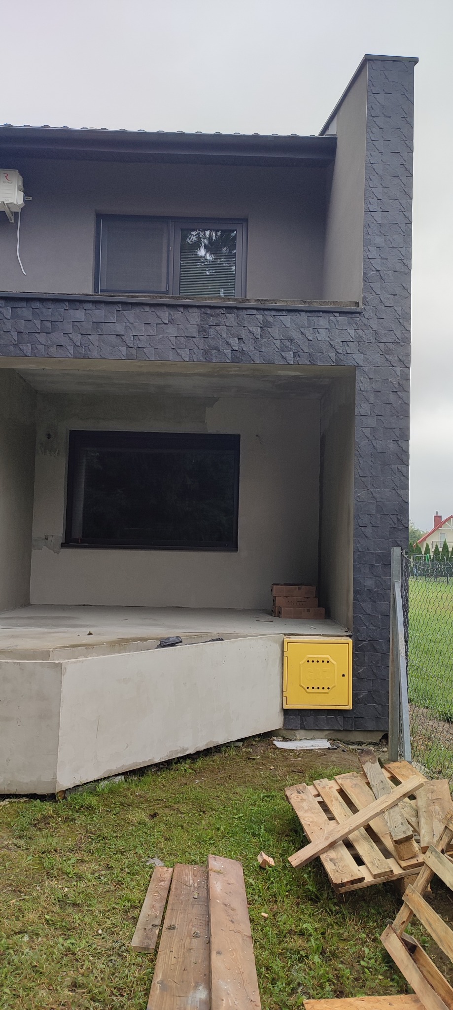 Surowy stan elewacji domu z szarą okładziną imitującą kamień, widoczna żółta skrzynka gazowa i palety drewniane na trawie.