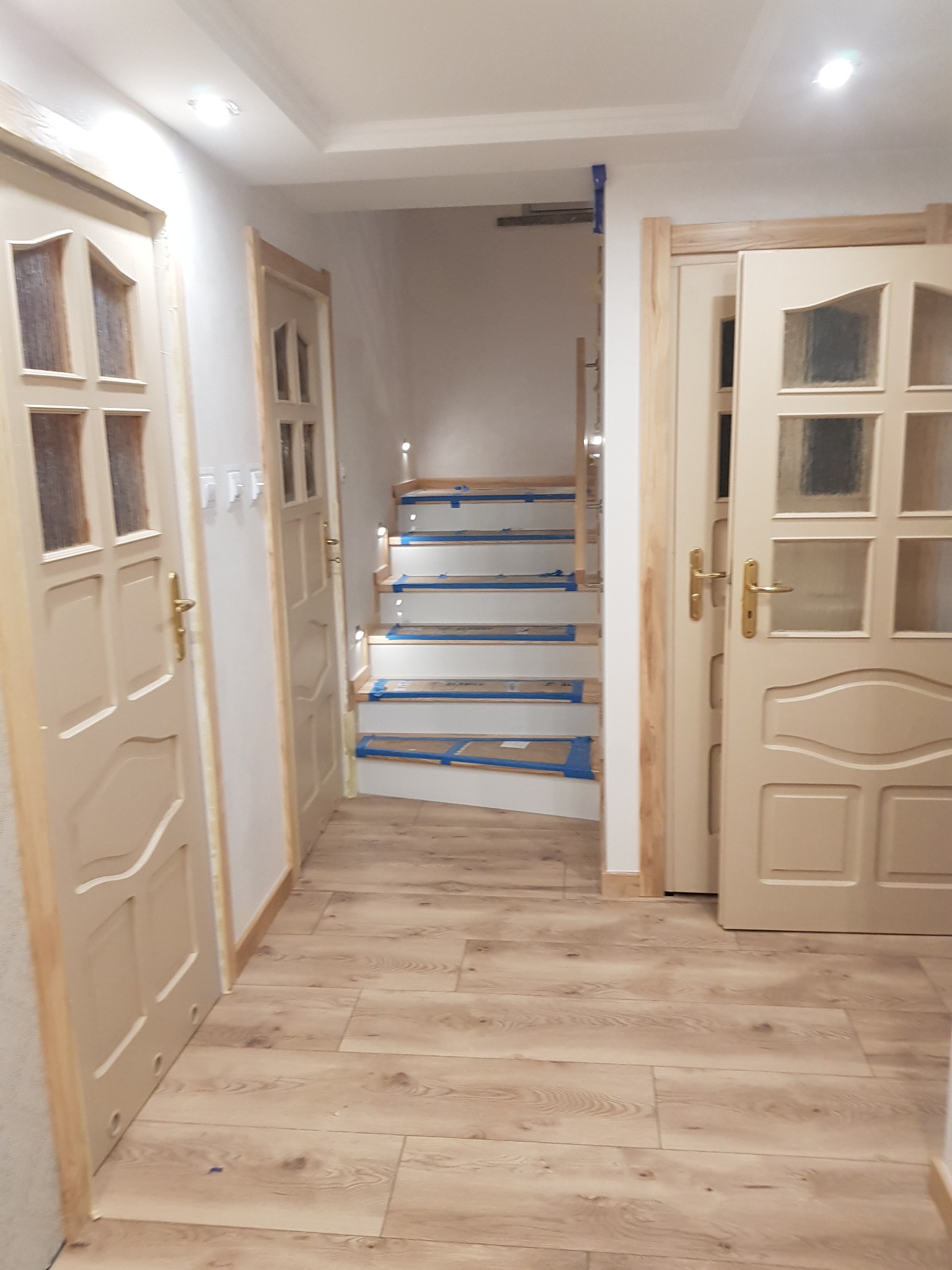 Wnętrze z dwiema beżowymi ościeżnicami drzwiowymi po bokach i schodami w głębi, zabezpieczonymi taśmą malarską, na podłodze panele imitujące drewno.
