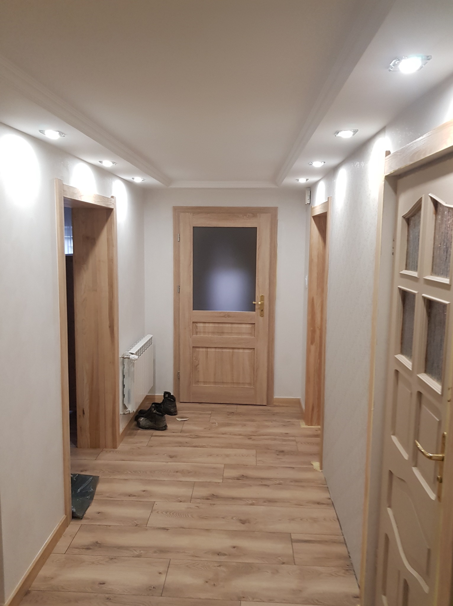 Korytarz po remoncie z trzema drewnianymi drzwiami, panelami podłogowymi w odcieniach brązu i beżu, oraz grzejnikiem pod oknem; na podłodze leżą buty i czarna folia zabezpieczająca.