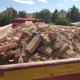 MV.finix Maciej Kusak - Kontener wypełniony gruzem ceglanym i betonowym, widoczny fragment żółto-czerwonego kontenera z logo KH-KIPPER, w tle zabudowa jednorodzinna i drzewa.