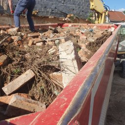 MV.finix Maciej Kusak - Pracownik stojący na stercie cegieł i gruzu w kontenerze, obok ramię koparki i fragment zdemontowanej ściany budynku z cegły.