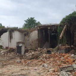 MV.finix Maciej Kusak - Resztki zniszczonego budynku z cegły i betonu, porośnięte roślinnością, z widocznym starym metalowym włazem.