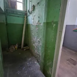 Wnętrze pomieszczenia z odpadającą zieloną farbą na ścianach, rurą pionową i oknem w górnej części ściany, widoczne ślady wcześniejszego użytkowania, z boku widoczne wejście do innego pomieszczenia.