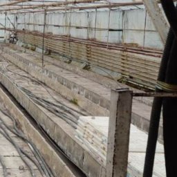 MV.finix Maciej Kusak - Wnętrze opuszczonej szklarni z metalową konstrukcją, betonowymi korytami i pozostałościami po uprawach. Widoczne rury i przewody. Na podłodze leżą resztki materiałów.