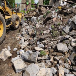 MV.finix Maciej Kusak - Żółta ładowarka kołowa częściowo widoczna obok sterty gruzu z cegieł i kamieni po rozbiórce budynku, z fragmentami zniszczonej konstrukcji w tle.