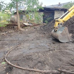 MV.finix Maciej Kusak - Żółta koparka Komatsu z łyżką nabiera ziemię i resztki po wycince drzew na placu budowy, w tle widoczny budynek w trakcie rozbiórki.