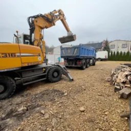 Żółta koparka Liebherr 316 ładuje gruz do niebieskiej wywrotki na placu budowy; w tle budynek mieszkalny i sterta drewna.