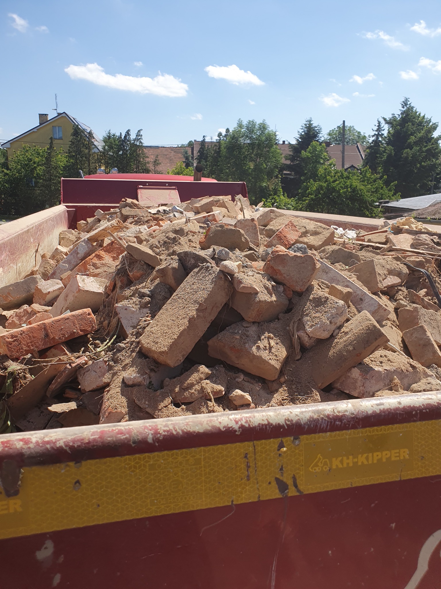 Kontener wypełniony gruzem ceglanym i betonowym, widoczny fragment żółto-czerwonego kontenera z logo KH-KIPPER, w tle zabudowa jednorodzinna i drzewa.
