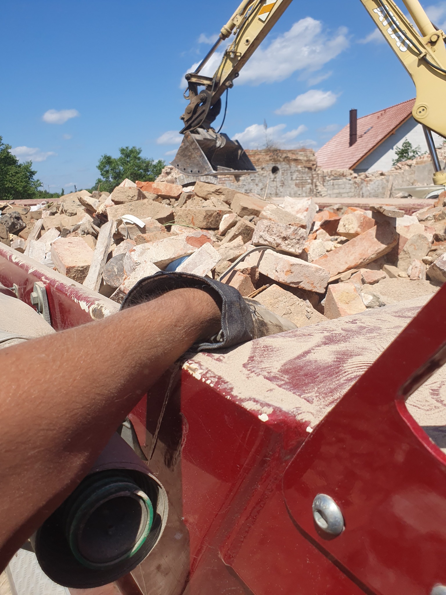 Ramię operatora opierające się o czerwoną krawędź maszyny z widokiem na stertę cegieł i gruzu po rozbiórce budynku, z żółtą koparką w tle i błękitnym niebem.