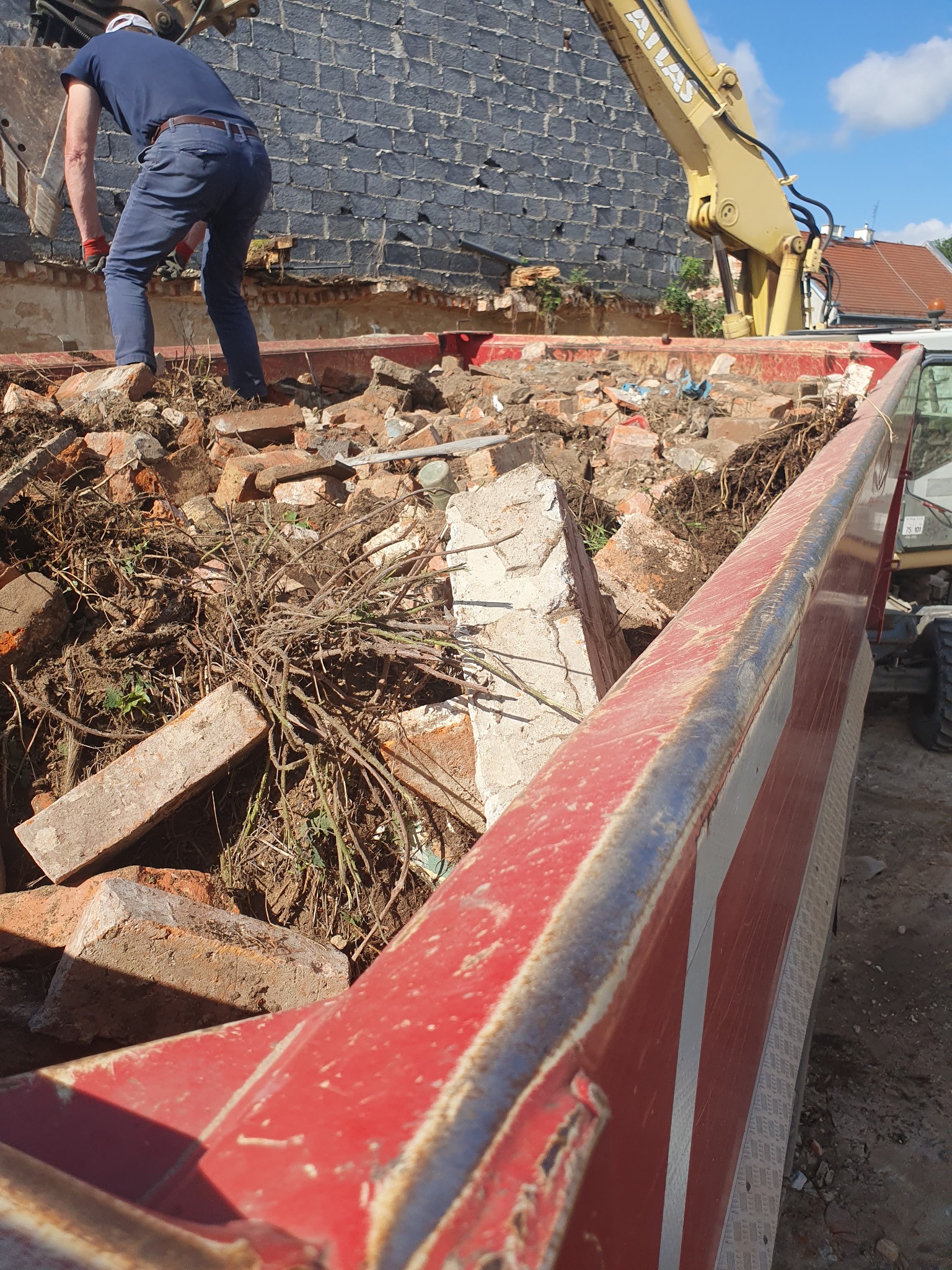 Pracownik stojący na stercie cegieł i gruzu w kontenerze, obok ramię koparki i fragment zdemontowanej ściany budynku z cegły.