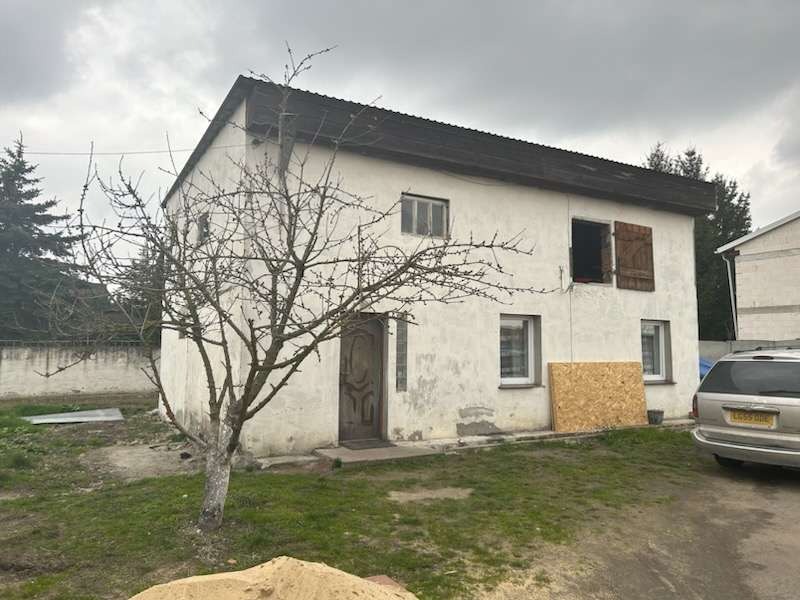 Dwukondygnacyjny budynek z częściowo odsłoniętym oknem na piętrze, z deską opartą o ścianę i stertą piasku na trawniku przed budynkiem.