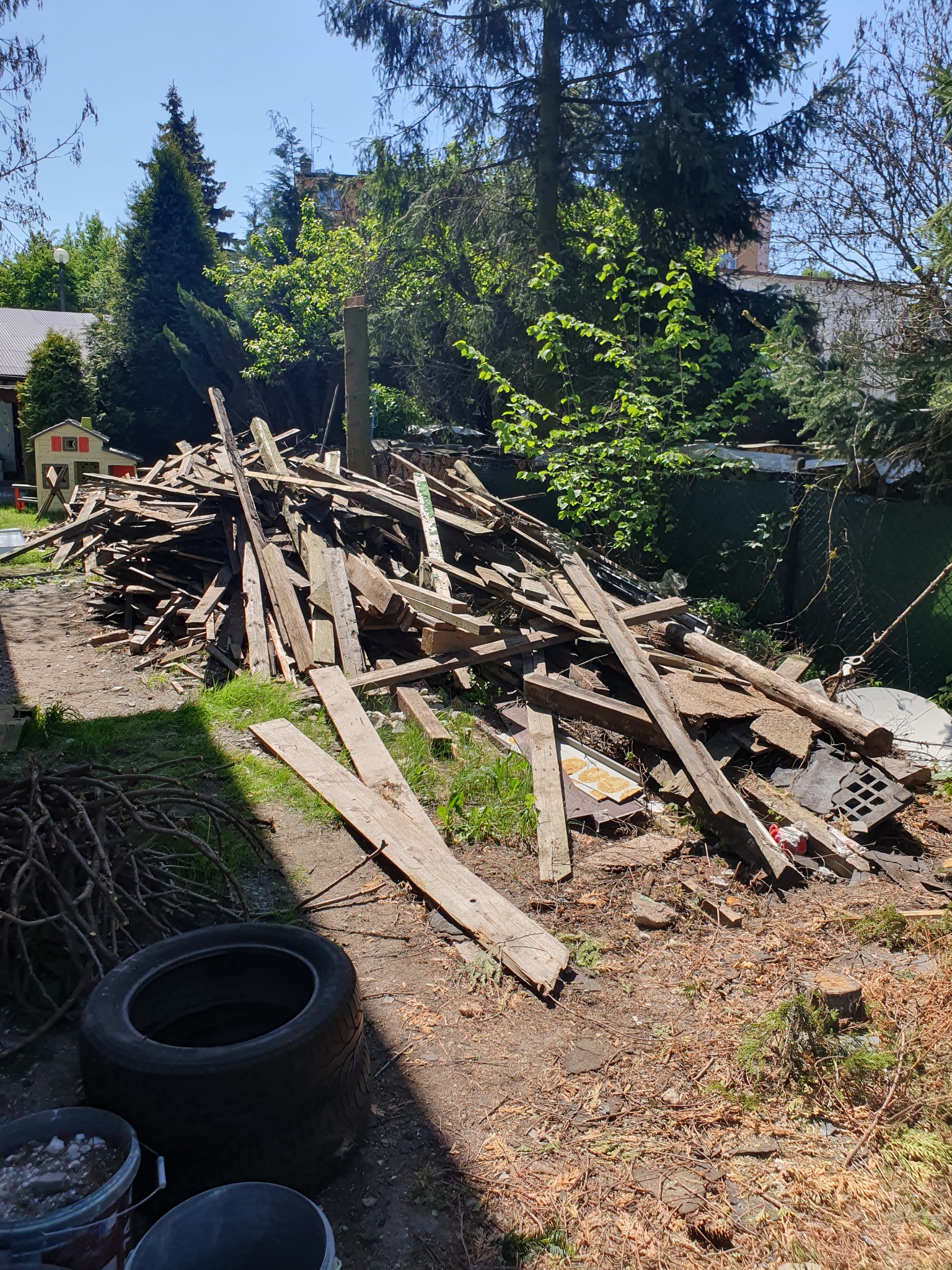 Sterta połamanych desek i innych odpadów budowlanych na ziemi w ogrodzie, w tle widoczne drzewa i budynki.