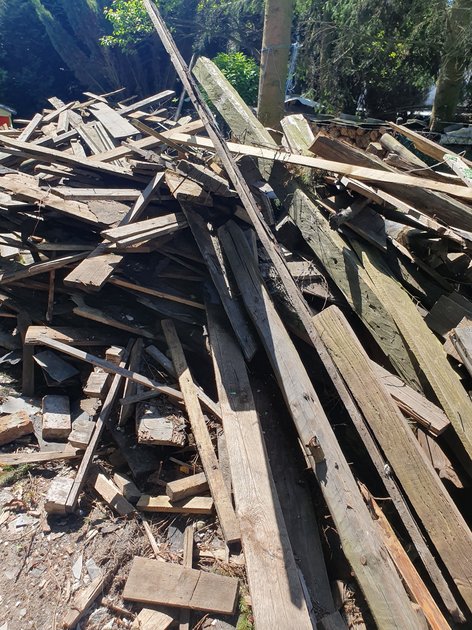 Sterta pociętego, starego drewna budowlanego z widocznymi gwoździami i śladami użytkowania, leżąca na ziemi w otoczeniu zieleni.