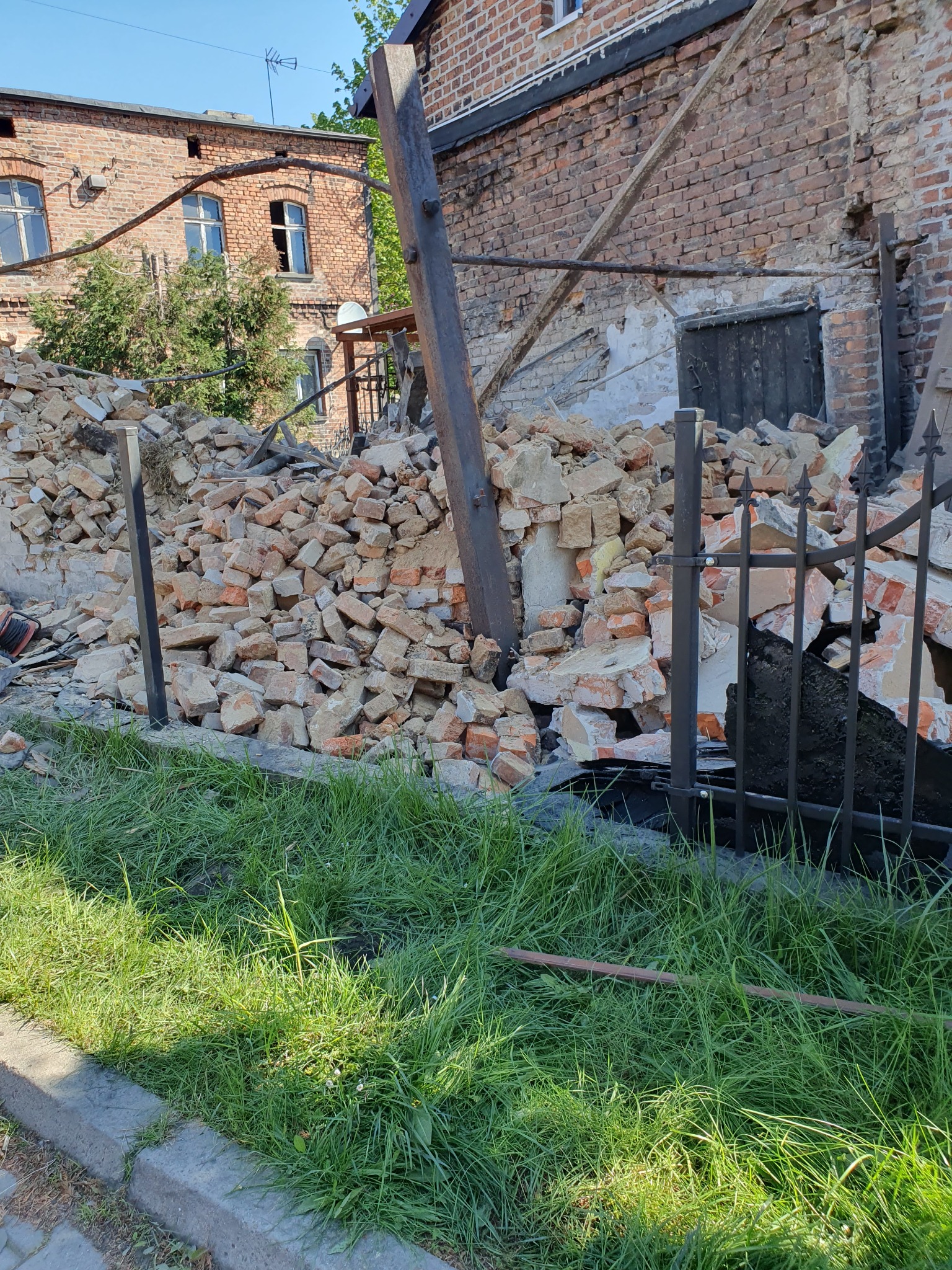 Sterta gruzu ceglanego po rozbiórce budynku, widoczna przez metalowe ogrodzenie, z fragmentami drewnianych belek podtrzymujących, zarośnięta trawą.