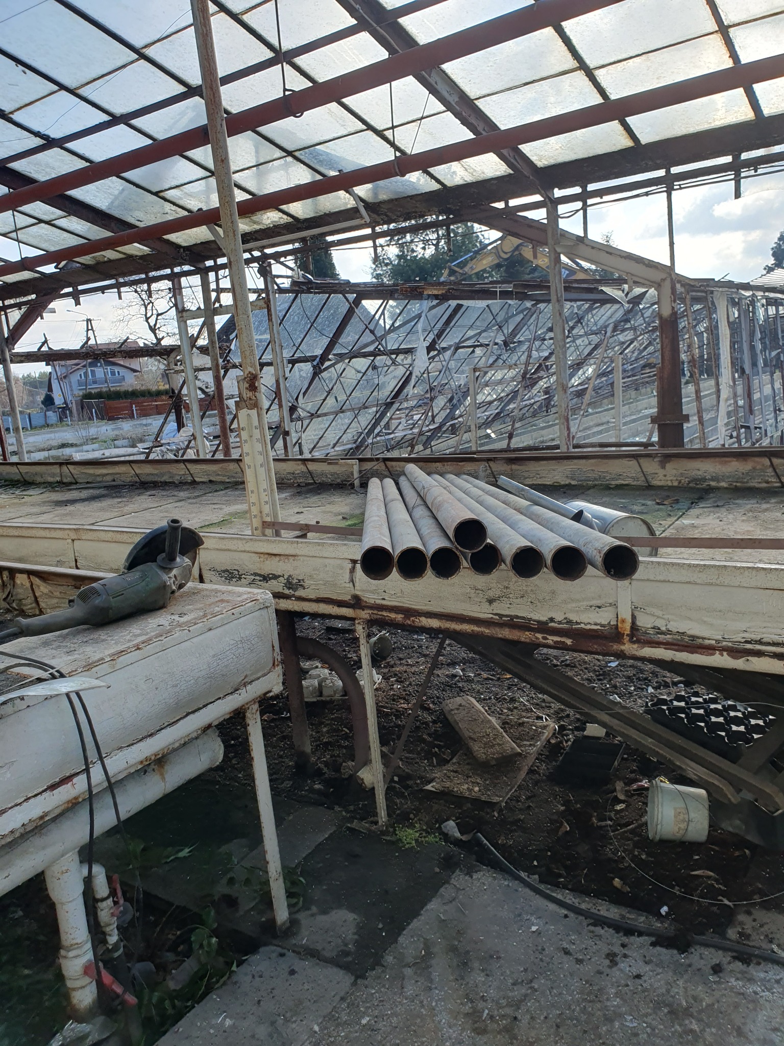 Wnętrze zrujnowanej szklarni z metalową konstrukcją i resztkami szyb, widoczne rury i szlifierka kątowa na stole roboczym.