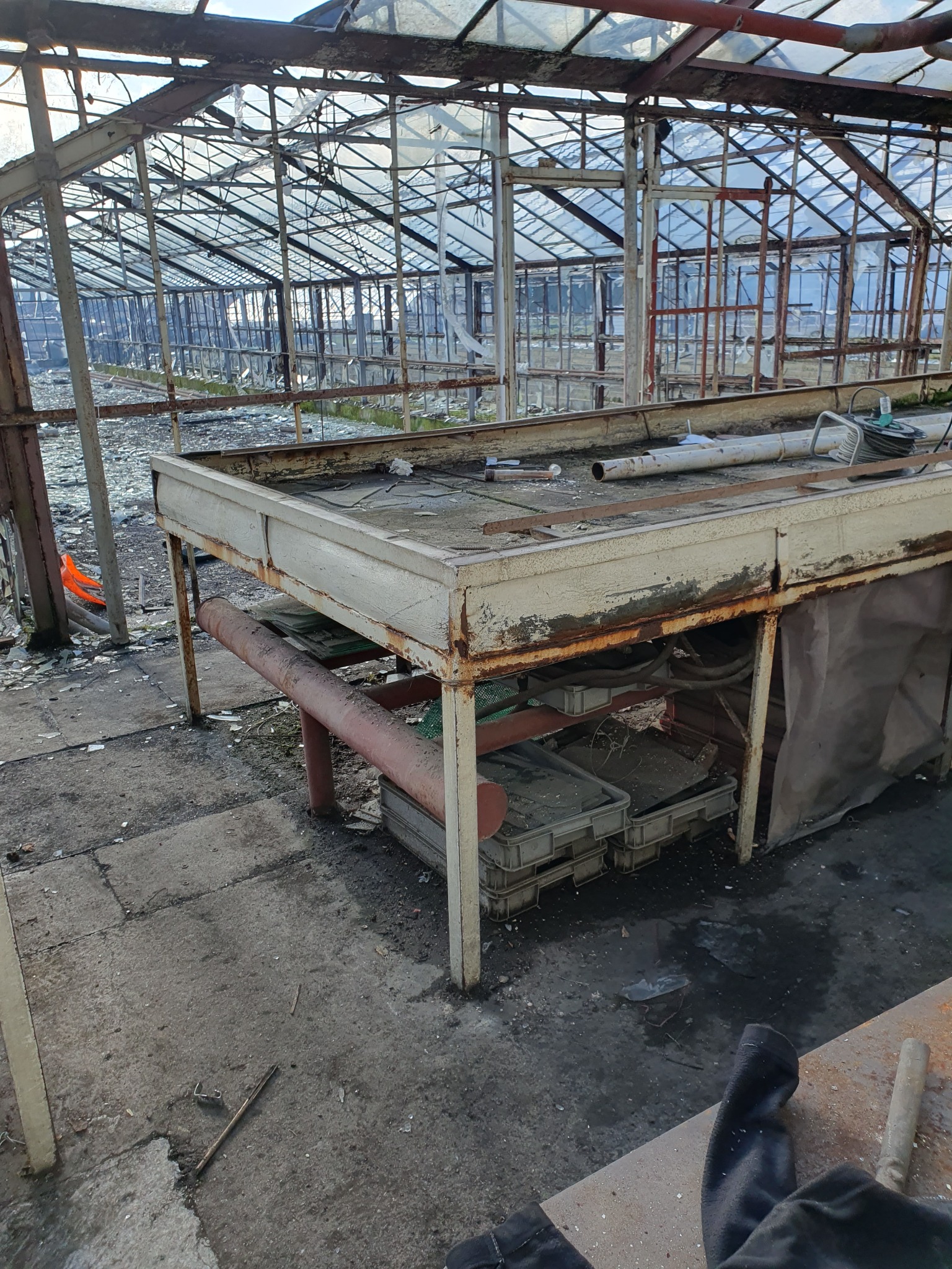 Wnętrze opuszczonej szklarni z metalową konstrukcją, rozbitymi szybami i zniszczonym stołem roboczym, widoczne ślady zaniedbania i rozkładu.