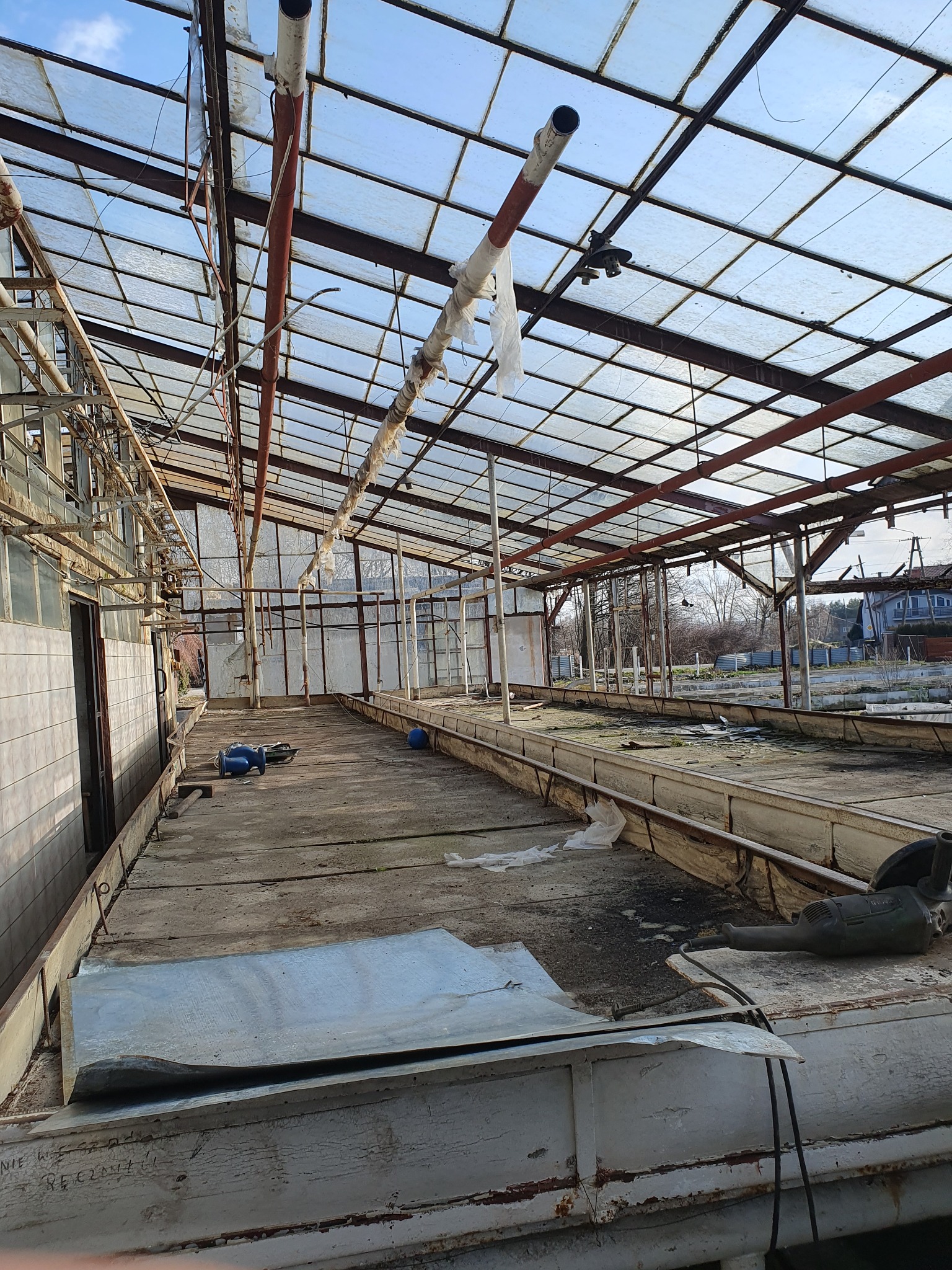 Wnętrze starej, zniszczonej szklarni w Sosnowcu, z widocznymi elementami konstrukcyjnymi, potłuczonymi szybami i narzędziami na podłodze, sugerujące przygotowania do rozbiórki lub opróżniania.