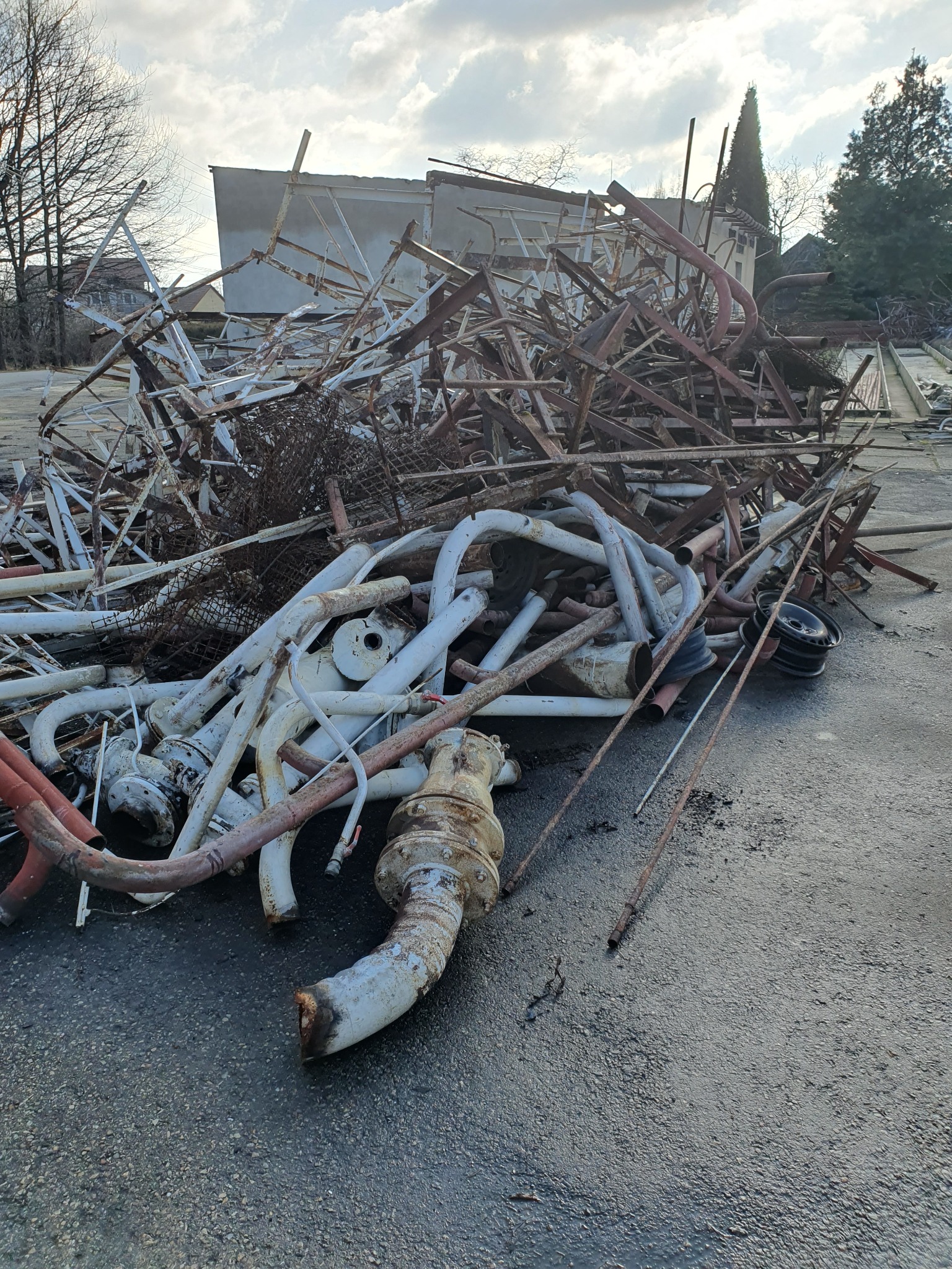 Sterta pordzewiałych metalowych rur i konstrukcji po wyburzeniu, z fragmentami budynku w tle i kołem samochodowym po prawej stronie