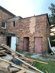 Fragment muru budynku z dwoma wejściami, z widocznymi śladami prac budowlanych i stertą desek na trawie.