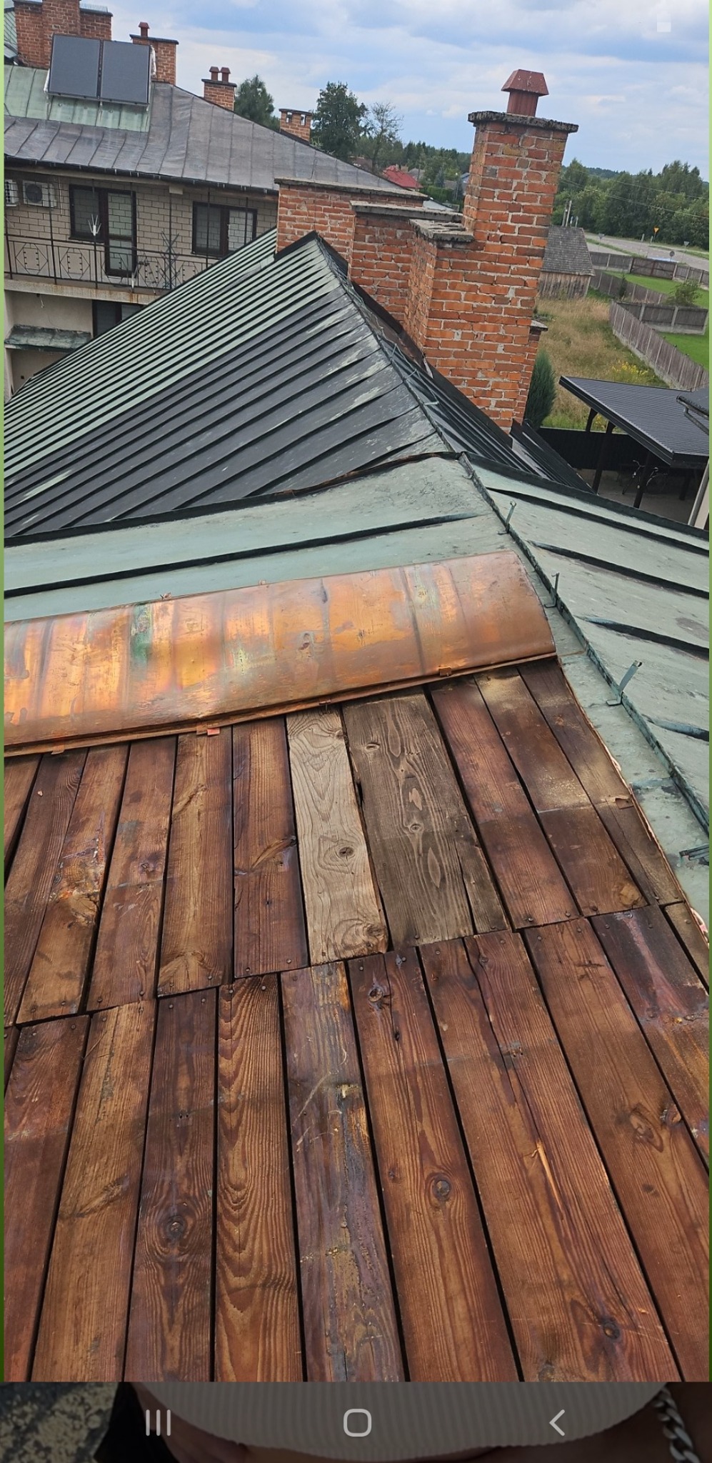 Fragment dachu z drewnianym wykończeniem, miedzianym elementem i metalową konstrukcją, widok z góry na tle zabudowań i zieleni.