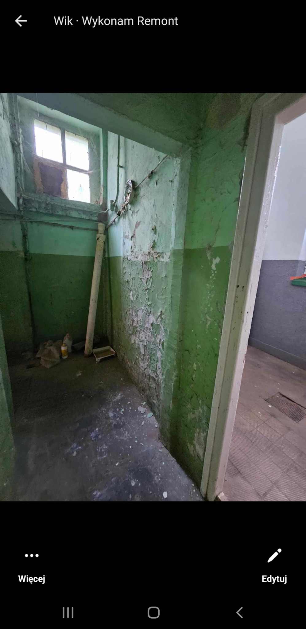 Wnętrze pomieszczenia z odpadającą zieloną farbą na ścianach, rurą pionową i oknem w górnej części ściany, widoczne ślady wcześniejszego użytkowania, z boku widoczne wejście do innego pomieszczenia.