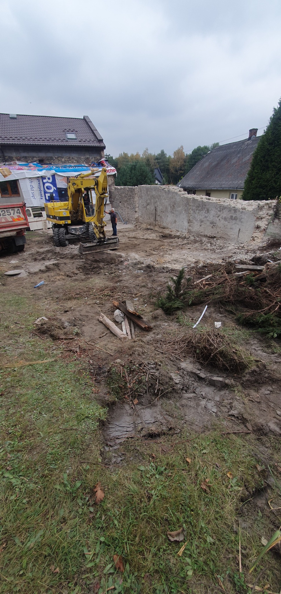 Żółta koparka przy częściowo rozebranej kamiennej ścianie budynku, widoczny gruz i pozostałości po roślinności.