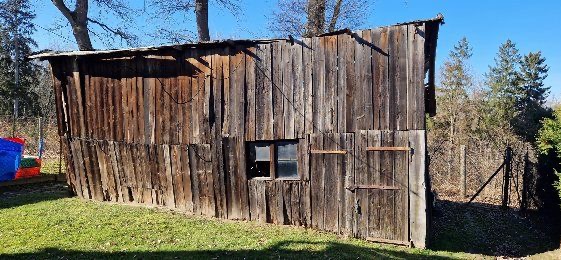 Stara, drewniana szopa z oknem i drzwiami na zewnątrz, z widoczną konstrukcją dachu i elementami zardzewiałego metalu, otoczona zielonym trawnikiem i drzewami w tle.