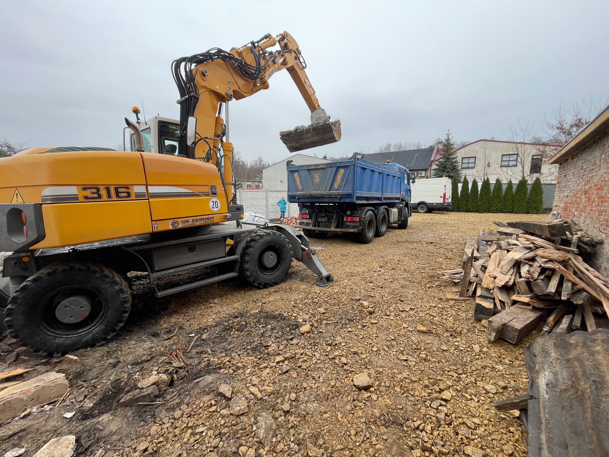Żółta koparka Liebherr 316 ładuje gruz do niebieskiej wywrotki na placu budowy; w tle budynek mieszkalny i sterta drewna.