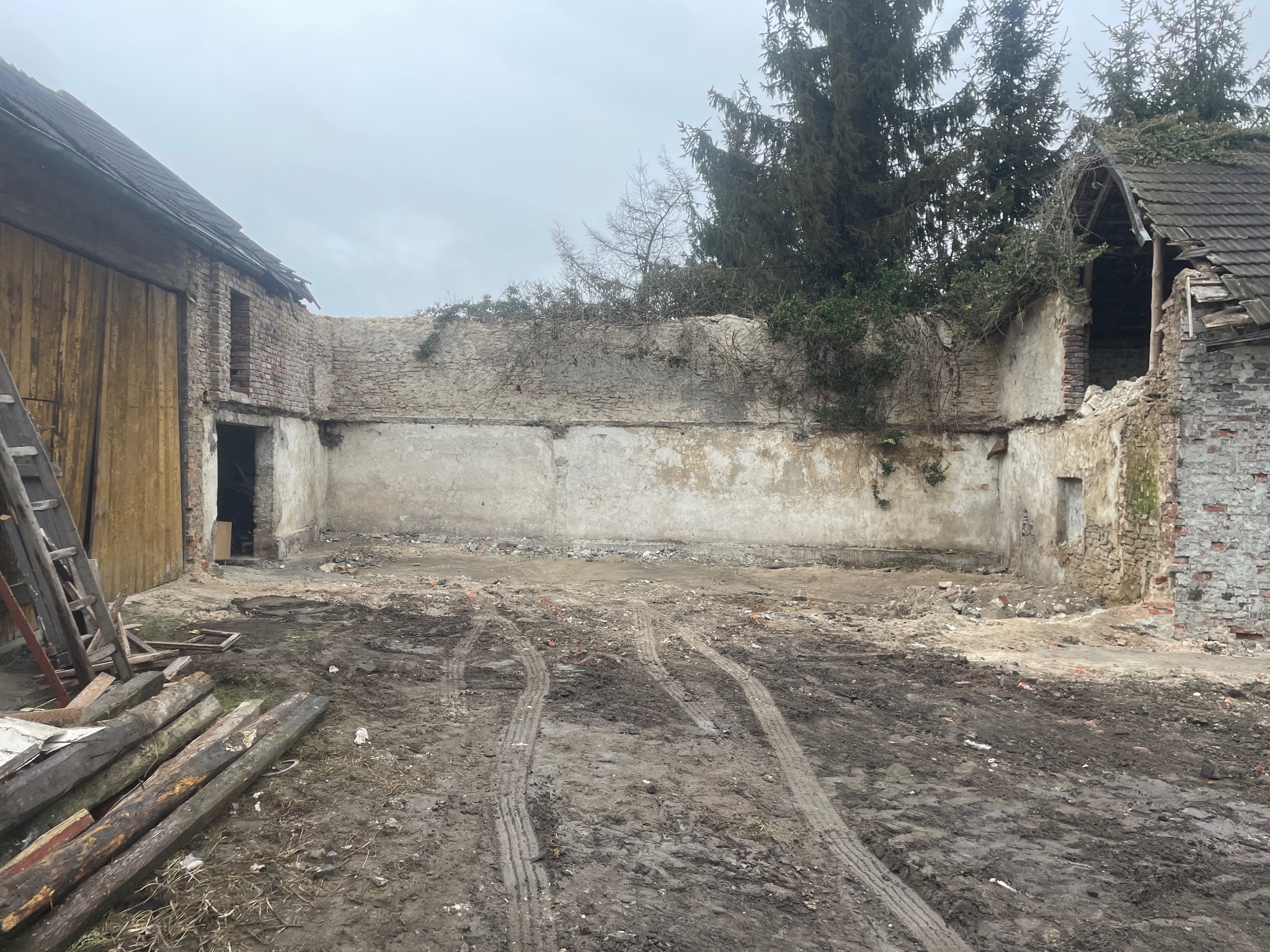 Częściowo rozebrany budynek gospodarczy z widocznymi śladami opon na błotnistym podłożu, pozostałości ścian z cegły i kamienia, oraz sterta drewnianych bali przy ścianie.