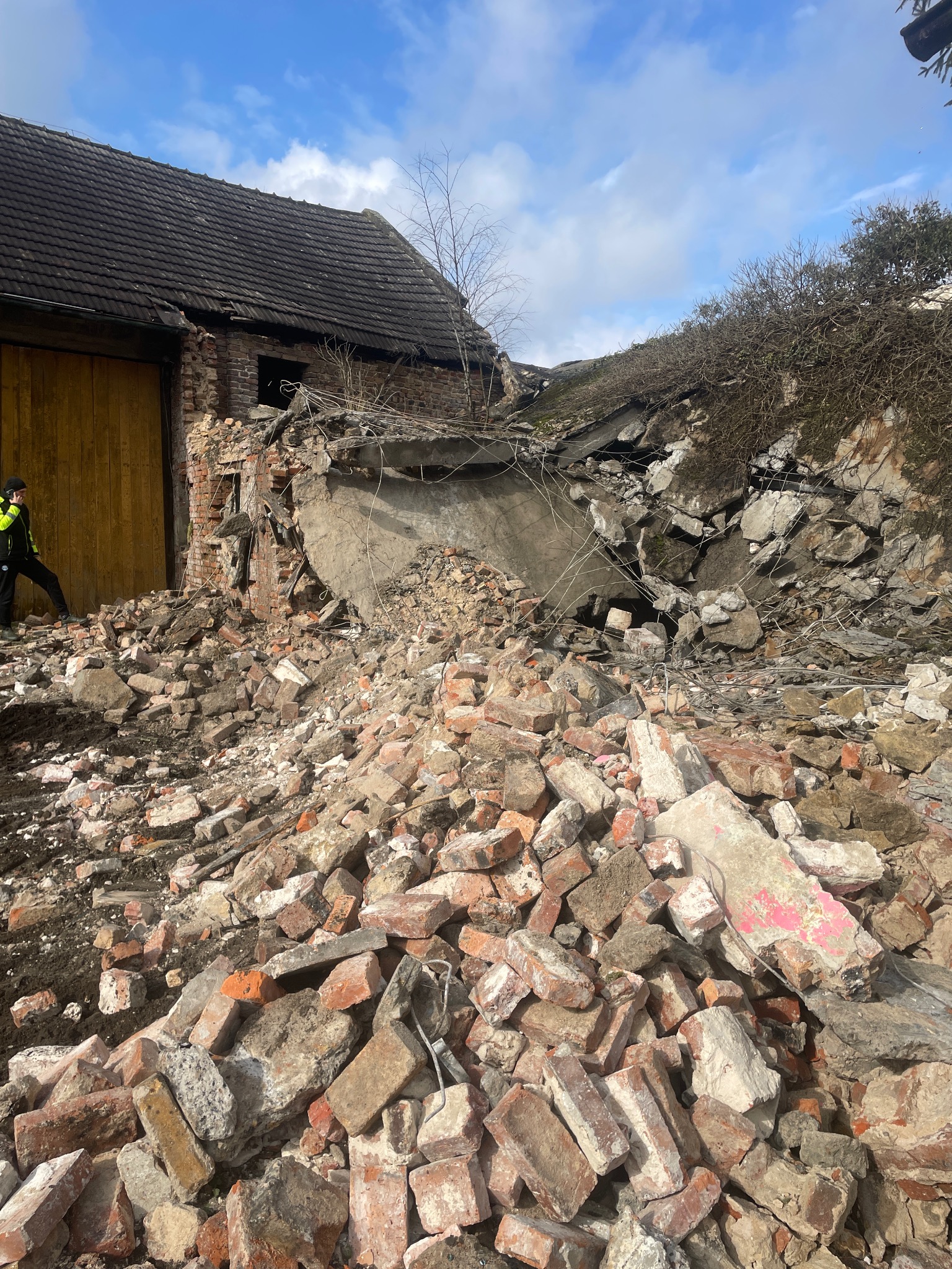 Stara, częściowo zawalona, ceglana stodoła z drewnianymi wrotami, z kupą gruzu i cegieł u podstawy, pracownik w odblaskowej kamizelce widoczny z boku.