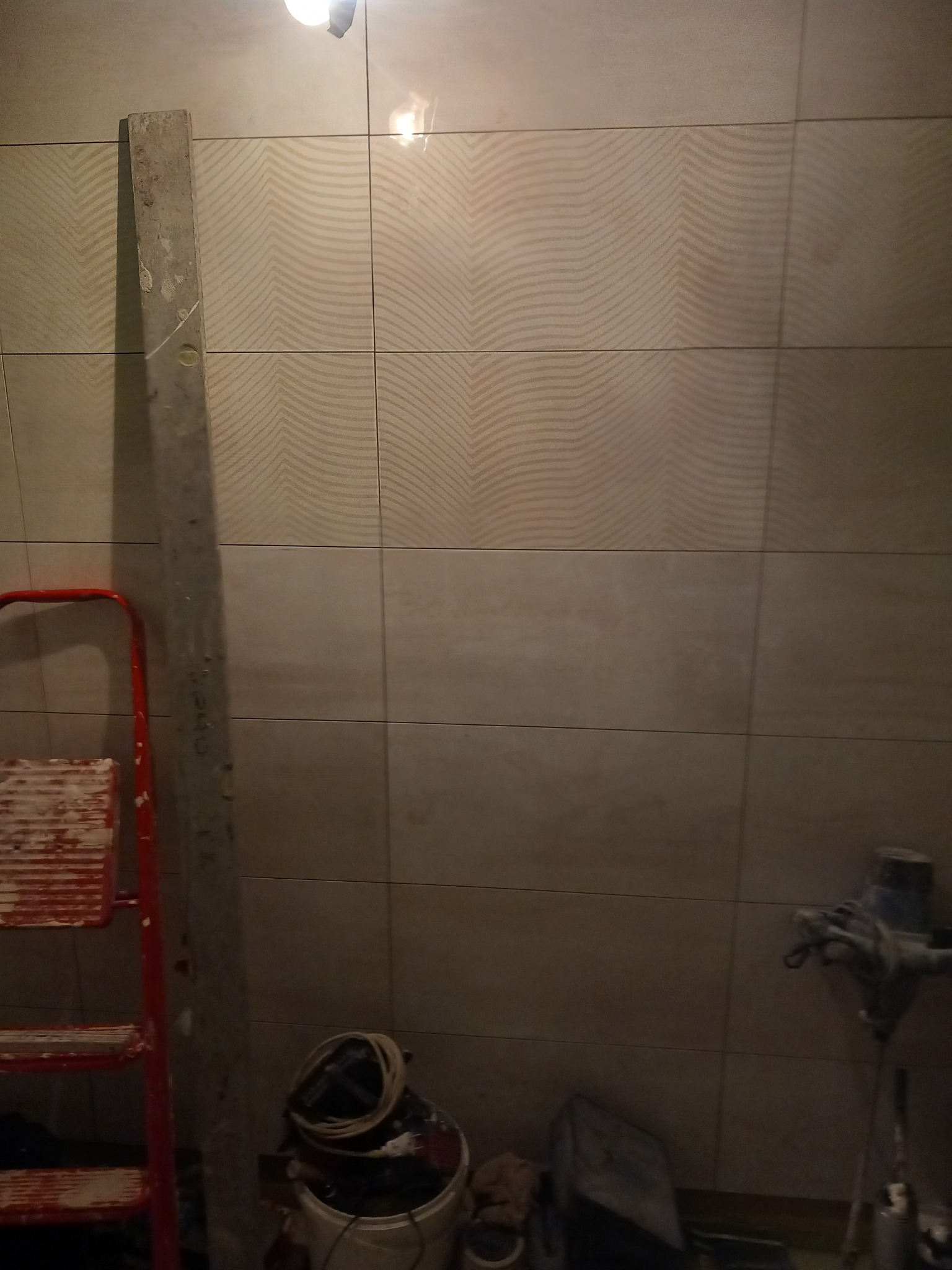 Ściana łazienki z ułożonymi płytkami o falistym wzorze, widoczna drabina malarska, poziomica i sprzęt do mieszania kleju, w trakcie prac wykończeniowych.